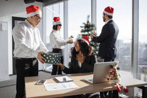 Asian and African american colleagues exchanging xmas gift box in festive office with christmas tree to celebrate winter holiday. People giving seasonal presents and feeling cheerful about Santa.