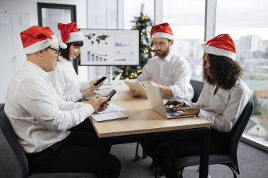 Büyük bir televizyon ekranı ve dekore edilmiş Noel ağacı ile konferans odasında, çeşitli etnik çeşitlilikteki iş adamları, masada işbirliği yapıyor, finansal tablolara ve cihazlara bakıyor..