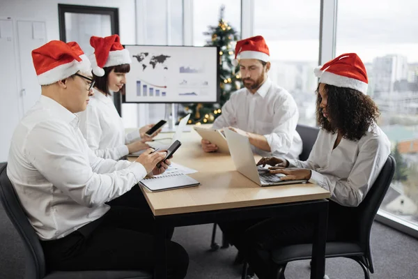 Festive diverse group of multiracial business people in conference room with big TV screen and decorated Christmas tree, cooperating at table, looking at papers with financial charts and gadgets.