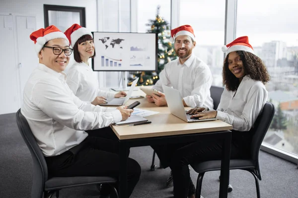 Festive diverse group of multiracial business people in conference room with big TV screen and decorated Christmas tree, cooperating at table, looking at papers with financial charts and gadgets.