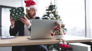 Masadaki Noel geyiğine odaklanın, Noel Baba şapkalı gülümseyen beyaz adamın bulanık arkaplanı çalışma masasında dizüstü bilgisayarla Noel ağacıyla süslenmiş bir şekilde otururken hediyesini açıyor..