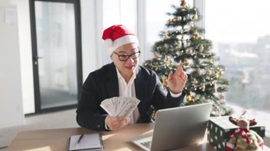 Noel Baba şapkalı yetişkin Asyalı zengin iş adamı para sayıyor, ofis işyerindeki gelir gelirlerini hesaplıyor Noel ağacıyla süslenmiş..