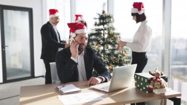 Takım elbiseli beyaz işadamı ve Noel Baba şapkası akıllı telefonla konuşurken taşınabilir dizüstü bilgisayar kullanarak Noel ağacını süsleyen meslektaşlarının arka planına bakıyor..