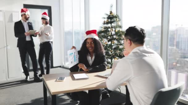 クリスマス タイム ジャケットとサンタの笑顔のアフリカのビジネスマンは オフィスとクリスマスツリーを飾っている同僚がアジアの従業員にグラフとチャートを持つ文書を示しています — ストック動画
