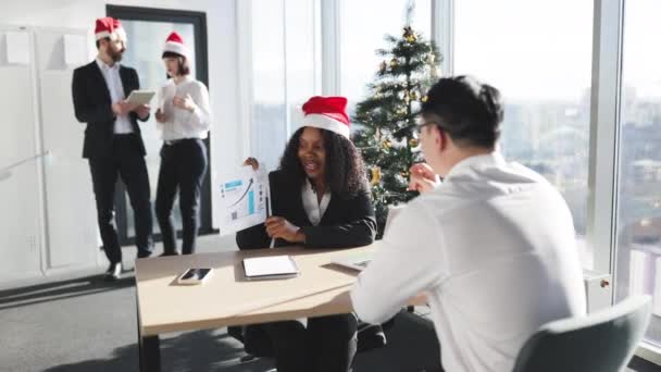 クリスマス タイム ジャケットとサンタの笑顔のアフリカのビジネスマンは オフィスとクリスマスツリーを飾っている同僚がアジアの従業員にグラフとチャートを持つ文書を示しています — ストック動画