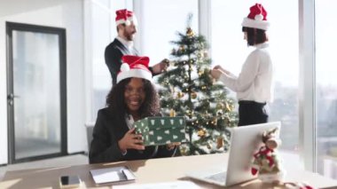 Noel Baba şapkalı başarılı bir yetişkin iş kadını elinde hediye kutusuyla masada otururken, meslektaşları modern ofiste Noel ağacını süslüyorlar..