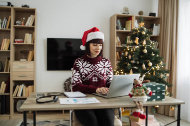 Kafkasyalı iş kadını Noel Baba şapkası takıyor Noel ağacının yanında oturuyor ve tatil sezonunda dizüstü bilgisayarda çalışıyor, bilgisayar klavyesinde daktilo kullanıyor..