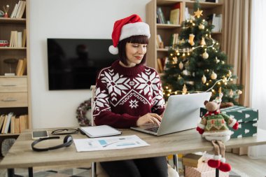 Kafkasyalı iş kadını Noel Baba şapkası takıyor Noel ağacının yanında oturuyor ve tatil sezonunda dizüstü bilgisayarda çalışıyor, bilgisayar klavyesinde daktilo kullanıyor..