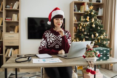 Noel Baba şapkalı beyaz iş kadını Noel ağacının yanındaki ofiste kameraya bakıyor ve tatil sezonu boyunca dizüstü bilgisayarda çalışıyor, klavyede daktilo yazıyor..