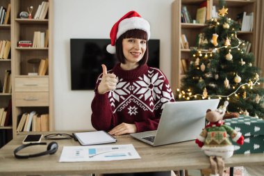 Noel Baba şapkalı beyaz iş kadını, Noel ağacının yanında otururken ve dizüstü bilgisayarda çalışırken bilgisayar klavyesinde yazarken kameraya bakıyor..