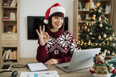 Noel Baba şapkalı beyaz iş kadını, Noel ağacının yanında otururken ve dizüstü bilgisayarda çalışırken bilgisayar klavyesinde yazarken kameraya bakıyor..