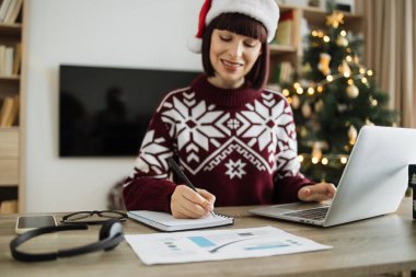 Noel Baba şapkalı, kalemli bir kadın. Tahta masadaki dizüstü bilgisayarın yanına yerleştirilen kağıt belgelere not alıyor. Beyaz tenli uzaktan kumandalı çalışanlar Noel ağacı süsleyerek evlerinden başlıyorlar..