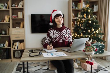Ofis masasında dijital cihazlarla poz veren güzel bir iş kadını portresi. Noel ağacıyla süslenmiş evde profesyonel faaliyetler yürüten serbest çalışan bir işçi..
