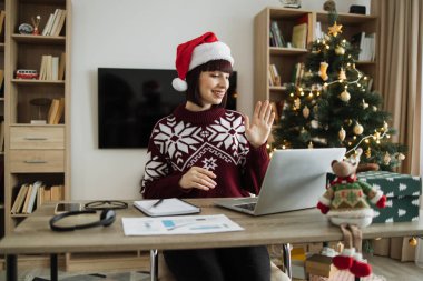 Ofisteki meslektaşını selamlarken, neşeli bir kadın yetişkin, taşınabilir bilgisayarın web kamerasında el sallıyor. Kafkas danışmanı noel ağacı yakınlarındaki teknolojileri kullanarak online konferans çağrısı alıyor.
