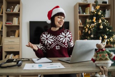 Noel Baba şapkalı mutlu bir kadın müşteriyle Noel 'de evde otururken görüntülü konuşma yapıyor. Neşeli işletme müdürü Noel ağacının arka planındaki projenin yeni detaylarını açıklıyor