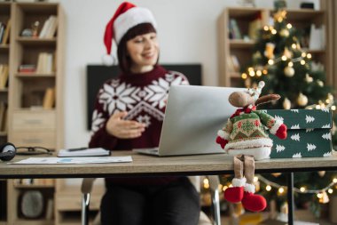 Masadaki Noel geyiğine odaklanın. Kafkasyalı, Noel şapkalı kadının bulanık arka planı. Çalışma masasında dizüstü bilgisayarla oturup Noel ağacıyla süslenmiş bir video görüşmesi..