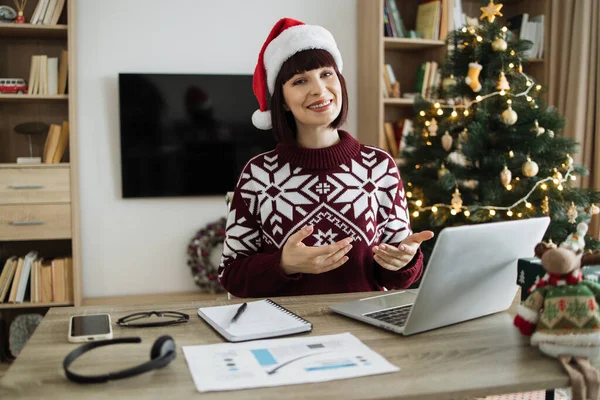 First person view of female adult posing to webcam while greeting colleague with New Year from home office. Caucasian consultant receiving online conference call using technologies near Christmas tree