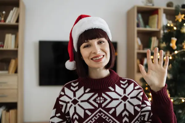 First person view of female waving hello while posing to webcam while greeting colleague or family with New Year from home on background of Christmas tree.
