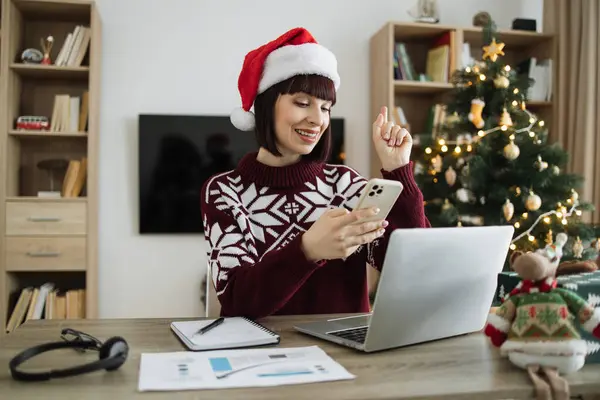 Noel Baba şapkalı olumlu bir kadın akıllı telefon tutuyor. Parmağını kaldır ve evde Noel ağacının arka planında çalışırken iyi bir fikir olduğuna dair işaret ver..