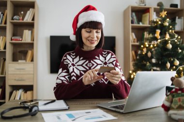 Noel Baba şapkalı çekici bir kadın elinde boş bir kredi kartı tutarken portatif bilgisayarda Noel ağacının arka planında yazı yazıyor. Kafkasyalı mutlu kadın evde çalışırken internet bankacılığı yapıyor.