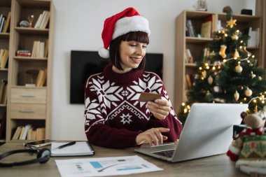 Noel Baba şapkalı çekici bir kadın elinde boş bir kredi kartı tutarken portatif bilgisayarda Noel ağacının arka planında yazı yazıyor. Kafkasyalı mutlu kadın evde çalışırken internet bankacılığı yapıyor.