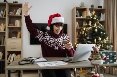 Noel Baba şapkalı mutlu bir kadın elinde boş bir kredi kartı olan dizüstü bilgisayara bakarken elini kaldırıyor. Mutlu Kafkasyalı yetişkin, Noel ağacının arka planında başarılı ödeme operasyonları yapıyor..