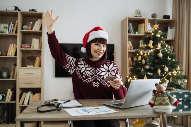 Noel Baba şapkalı mutlu bir kadın elinde boş bir kredi kartı olan dizüstü bilgisayara bakarken elini kaldırıyor. Mutlu Kafkasyalı yetişkin, Noel ağacının arka planında başarılı ödeme operasyonları yapıyor..