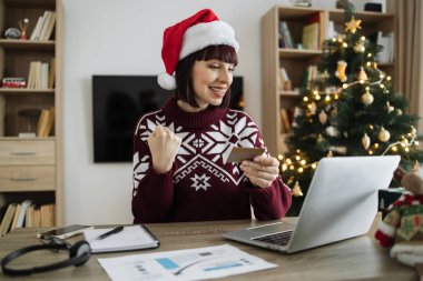 Noel Baba şapkalı heyecanlı genç bir kadın elinde boş bir kredi kartıyla dizüstü bilgisayar ekranına bakarken yumruk kaldırıyor. Mutlu Kafkasyalı yetişkin, Noel ağacının arka planında başarılı ödeme operasyonları yapıyor..
