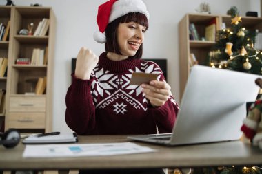 Noel Baba şapkalı heyecanlı genç bir kadın elinde boş bir kredi kartıyla dizüstü bilgisayar ekranına bakarken yumruk kaldırıyor. Mutlu Kafkasyalı yetişkin, Noel ağacının arka planında başarılı ödeme operasyonları yapıyor..