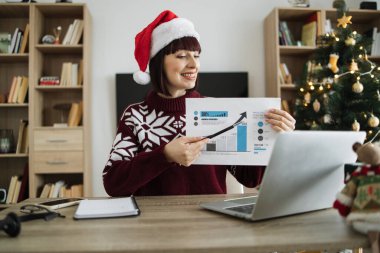 Noel Baba şapkalı kendine güvenen yetişkin iş kadını, Noel ağacıyla süslenmiş online sunum yaparken grafik materyallere işaret ediyor..