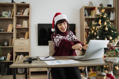 Noel Baba şapkalı başarılı bir yetişkin kadın ve sıcak kış kıyafetleri Noel ağacıyla süslenmiş oturma odasında bilgisayar ekranına bakarken heyecandan dans ediyor..