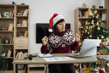 Noel Baba şapkalı başarılı bir yetişkin kadın ve sıcak kış kıyafetleri Noel ağacıyla süslenmiş oturma odasında bilgisayar ekranına bakarken heyecandan dans ediyor..