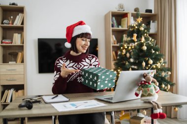 Noel Baba şapkalı gülümseyen beyaz kadın dizüstü bilgisayarla masada otururken hediyesini açıyor. Mutlu kadın kutusunu açıyor ve Noel ağacıyla süslenmiş evde uzaktan çalışma sırasında heyecanlanıyor..