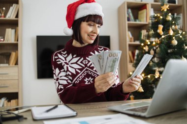 Noel Baba şapkalı, genç, heyecanlı, beyaz bir kadının ev ofisinde Noel ağacıyla süslenmiş paraları sayarak el ele tutuşması. Muhasebe, vergi ve finans kavramı.