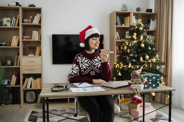 Portrait of attractive woman freelancer in Santa hat and casual wear recording audio message, sound voice on modern phone while sitting at table with gadgets and papers on background of Christmas tree