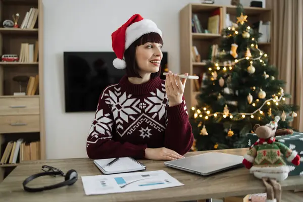 Portrait of attractive woman freelancer in Santa hat and casual wear recording audio message, sound voice on modern phone while sitting at table with gadgets and papers on background of Christmas tree