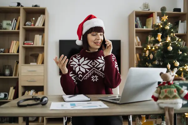 Side view of business person in Santa hat typing on laptop during call on cell phone in Christmas decorated room. Professional caucasian manager discussing report while looking at screen during call.