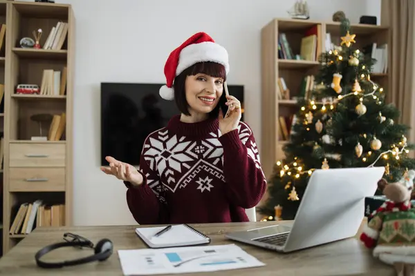 Side view of business person in Santa hat typing on laptop during call on cell phone in Christmas decorated room. Professional caucasian manager discussing report while looking camera.