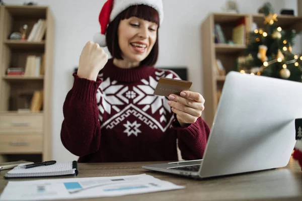Excited young woman in Santa hat raising fist while looking at laptop screen with blank credit card in hand. Happy caucasian adult making successful payment operations on background of Christmas tree.