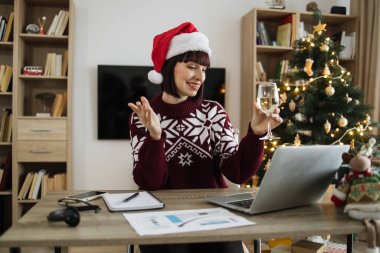 Noel Baba şapkalı, elinde şampanya bardağıyla, video görüşmesinde müşteriyle konuşurken Noel ağacının arka planında oturan mutlu beyaz kadın yönetici..
