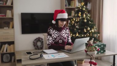 Kafkasyalı iş kadını Noel Baba şapkası takıyor Noel ağacının yanında oturuyor ve tatil sezonunda dizüstü bilgisayarda çalışıyor, bilgisayar klavyesinde daktilo kullanıyor..