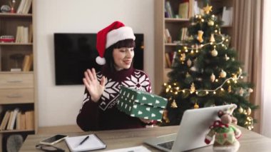 Noel Baba şapkalı gülümseyen beyaz kadın dizüstü bilgisayarla masada otururken hediyesini açıyor. Mutlu kadın kutusunu açıyor ve Noel ağacıyla süslenmiş evde uzaktan çalışma sırasında heyecanlanıyor..