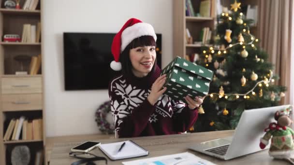 头戴圣诞礼帽的多愁善感的成年女子打开礼物 一边举手表决 一边看着家里装饰着圣诞树的相机 心情愉快的高加索女性成功购买了冬眠用品 — 图库视频影像