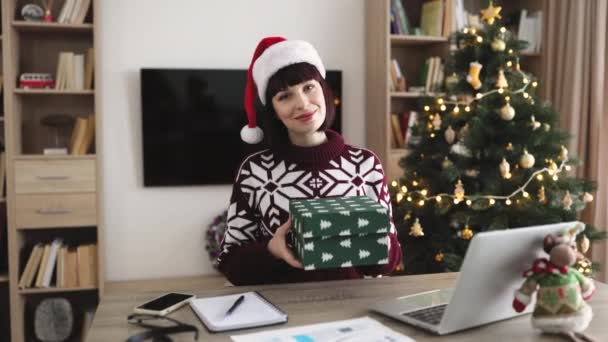 サンタの帽子とカジュアルな服で幸せな白人ビジネスマンは カメラを見ているクリスマスツリーで飾られた自宅で机に座っている間 オンライン注文でお祝いのボール紙箱を提供しています — ストック動画