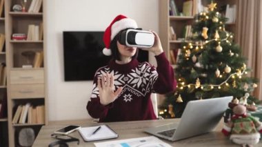 Kafkasyalı genç iş kadını Noel Baba şapkası takıyor VR gözlükler takıyor havaya dokunuyor ve el kol hareketleri yapıyor, Noel zamanı evde Noel ağacının yanında aletlerle oturuyor..