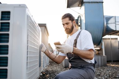 Odaklanmış sakallı usta dijital çoklu metre tutan eldivenler giyiyor ve klimanın voltaj seviyesini kontrol ediyor. Beyaz adam açık terasta fabrika ekipmanlarıyla çalışıyor..
