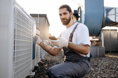 Odaklanmış sakallı usta dijital çoklu metre tutan eldivenler giyiyor ve klimanın voltaj seviyesini kontrol ediyor. Beyaz adam açık terasta fabrika ekipmanlarıyla çalışıyor..