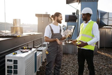 Beyaz miğferli ve yansıtıcı yelekli Afro-Amerikan mühendis Kafkasyalı tamircinin çalışmalarını değerlendirirken klimaya bakıyor. Bitki ekipmanları ve bakım konsepti.