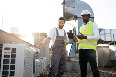 Beyaz miğferli ve yansıtıcı yelekli Afro-Amerikan mühendis Kafkasyalı tamircinin çalışmalarını değerlendirirken klimaya bakıyor. Bitki ekipmanları ve bakım konsepti.