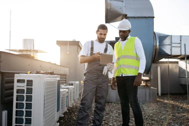 Afrika kökenli Amerikalı yönetici ve beyaz teknisyen yan yana duruyor ve modern tablet ekranlara bakıyor. Nitelikli fabrika işçileri çatıdaki klimalar hakkında bilgi okuyorlar.